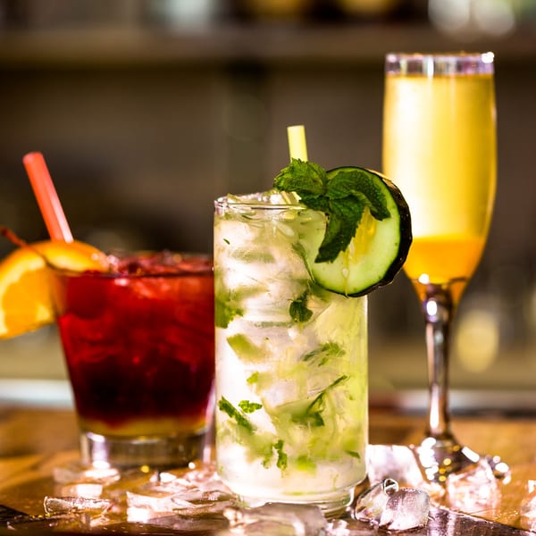 Three vibrant cocktails on a bar, including a red drink, a mojito, and a yellow mimosa.