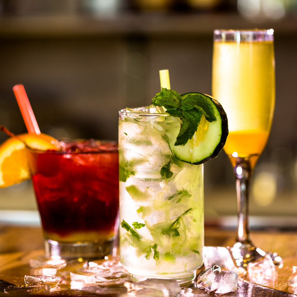Three vibrant cocktails on a bar, including a red drink, a mojito, and a yellow mimosa.