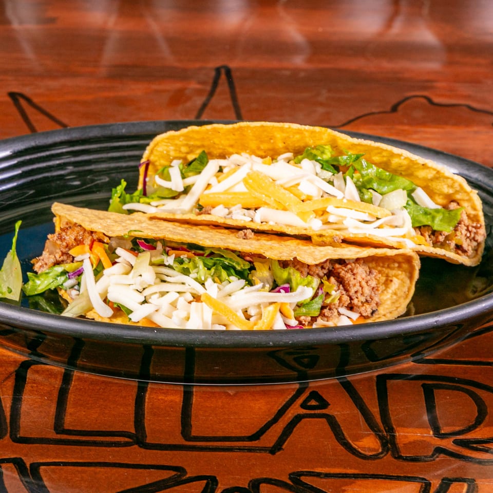 Two crispy tacos filled with seasoned ground beef, shredded lettuce, and mixed cheese on a black plate.