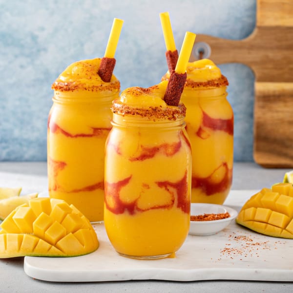 Three mango chamoyadas in mason jars with chili-lime straws and fresh mango on a white board.
